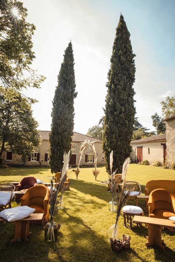An outdoor ceremony or cocktail hour setup on a lawn in front of a French country estate with stone and rendered facades, terracotta roof tiles, and burgundy shutters. The seating arrangement features mustard yellow and burgundy velvet armchairs paired with low wooden coffee tables, styled in a boho-rustic theme. Large glass vase bottles filled with dried pampas grass and dried grasses line the central aisle, and a macramé arch adorned with pampas grass marks the focal point between two tall cypress trees. The wide shot captures the full layout of the space with the venue buildings visible in the background. Potential venue feature image.
