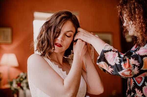 A getting-ready moment captured indoors in a warm terracotta-walled room, where a woman with curly hair wearing a dark floral kimono-style robe helps the bride put on or adjust an earring. The bride has a short wavy bob, bold red lipstick, and a small arm tattoo, and is wearing a white lace-trimmed bridal gown or slip. A table lamp and what appears to be a floral arrangement are softly lit in the background. The composition is a close-up portrait shot with warm ambient lighting emphasizing the intimate, candid nature of the moment.