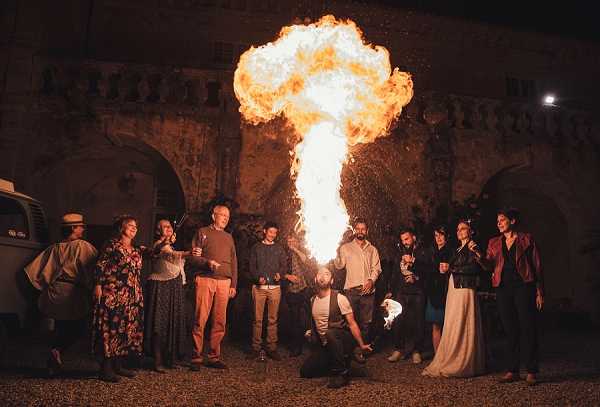A fire performer crouches low and blows a large column of flame into the night air during what appears to be an outdoor wedding reception entertainment segment. Approximately twelve guests stand in a semicircle watching the performance, dressed in a mix of casual and semi-formal attire including a floral maxi dress, burgundy leather jacket, and earth-toned separates. The setting is the exterior courtyard of a stone chateau or manor house, with arched architectural details visible in the background, and a vintage white van parked to the left. The wide-group shot is lit almost entirely by the dramatic burst of fire, casting warm orange light across the guests' faces against a dark night sky.