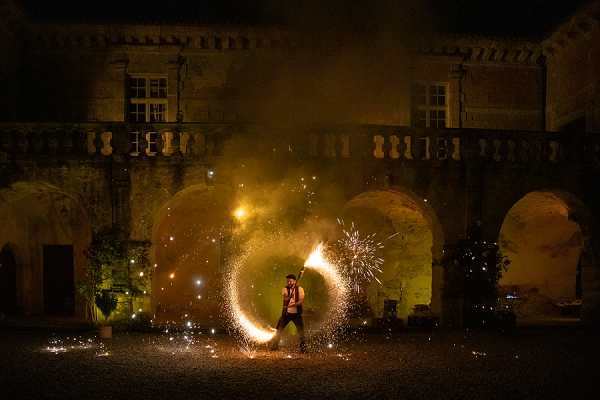A fire performer is captured mid-act during a nighttime wedding entertainment segment, spinning fire poi or a fire staff to create a large circular trail of sparks and flames. The performance takes place in the courtyard of a French chateau, with stone arched colonnades and a balustrade upper-level facade illuminated in warm golden-green uplighting. The long-exposure wide shot captures the full arc of the fire trail surrounding the performer, with sparks scattering across the gravel courtyard floor. The overall color palette contrasts deep blue-black night sky against the warm orange and gold of the fire and the green-tinted floodlit stone architecture. Potential venue feature image.