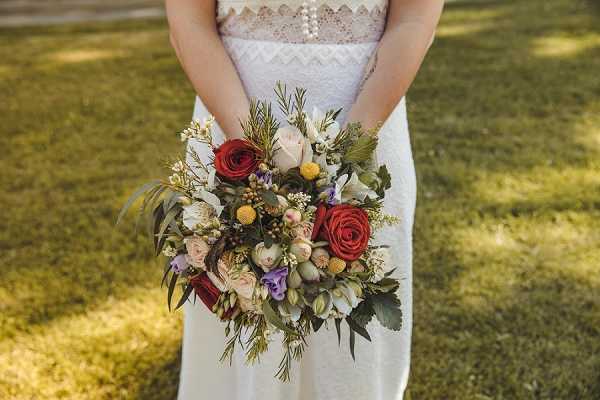 Close-up portrait of a bride holding a large, loosely arranged bouquet in front of her. The bride is wearing a white lace dress with a geometric chevron bodice detail and pearl embellishments at the waist. The bouquet features deep red roses, blush pink roses, lavender lisianthus, white blooms, yellow craspedia (billy balls), berry clusters, and abundant greenery including rosemary sprigs and eucalyptus leaves, styled in a wild, garden-gathered boho aesthetic. The shot is cropped from chest to mid-thigh, keeping focus entirely on the dress detail and bouquet against a lawn background.