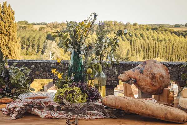 A close-up detail shot of an outdoor rustic wedding reception table styled with a harvest-inspired food display, set on a stone terrace overlooking a rolling wooded landscape. The table features a whole cured ham on the bone, a baguette, fresh lettuces, a decorative plate, and a floral textile runner with a warm earthy pattern. Two green glass bottles hold an arrangement of wildflowers, eucalyptus, yellow blooms, and purple sprig flowers in an informal, foraged style. The overall decor palette combines deep greens, mustard yellows, and warm terracotta tones, consistent with a rustic or countryside-themed wedding aesthetic.