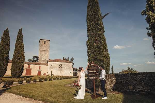 A bride and groom pose together next to a wooden directional wedding sign in the grounds of a French stone chapel or church with a square tower, surrounded by tall cypress trees and manicured hedges. The bride wears a long white gown and holds a small bouquet with deep red/burgundy tones, while the groom is dressed in dark navy trousers and a light shirt with suspenders. The wooden sign features calligraphy text in French indicating event directions including 'Cérémonie,' 'Cocktail,' and 'Dîner.' The shot is a wide portrait taken outdoors on a sunny day, capturing both the couple and the historic stone venue building in the background. Potential venue feature image.