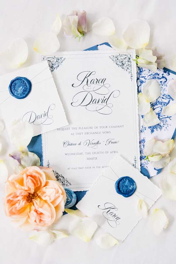 A flat lay detail shot of a wedding invitation suite for Karen and David, set against a white background scattered with cream and pale lavender flower petals and a peach garden rose. The main invitation card features calligraphy script lettering with ornate black corner flourishes, listing the venue as Château de Versailles, France, on Wednesday the eighth of April MMXIX. The suite includes two small place cards — one reading 'David' and one reading 'Karen' — each sealed with a blue wax seal, alongside a blue and white toile-patterned card and a deep navy blue silk ribbon. The overall stationery palette is white, black, and navy blue, with a classic French-inspired design aesthetic.