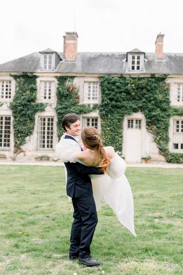 A couple portrait shot outdoors on the lawn of a French chateau, with the groom lifting the bride and both facing each other closely. The groom wears a navy suit, and the bride wears a white gown with a flowing skirt and has her auburn hair styled in a half-up updo with a small gold hair accessory. The ivy-covered stone chateau facade with mansard roof, brick chimneys, and multi-pane windows fills the background, rendered softly out of focus. The image is a medium portrait composition with a bright, airy, film-inspired aesthetic. Potential venue feature image.