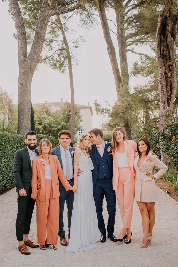 A group portrait of seven people posed outdoors on a gravel path, with a chateau building visible in the background among tall pine trees. The bride wears a flowing white pleated dress with a floral crown, and the groom is dressed in a navy three-piece suit with a blush pink bow tie and a floral boutonnière. Two female guests wear coordinating coral/salmon trouser suits, one male guest wears a dark green suit, and another female guest wears a blush beige blazer and shorts. The group is loosely arranged, with the couple at the center gazing at each other. The overall color palette of the wedding party leans toward coral, blush, and navy tones, suggesting a boho-romantic style. This is a full-length portrait shot taken in natural outdoor light.