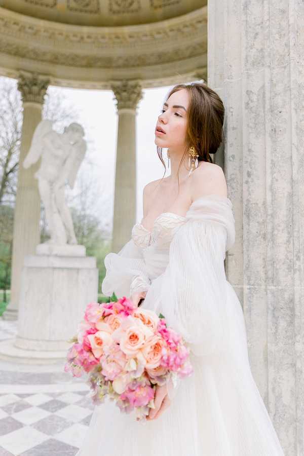 A bridal portrait of a single bride leaning against a stone column within a classical open rotunda or temple structure featuring Corinthian columns, a domed ceiling, and a white marble statue on a pedestal in the background. The bride wears an off-the-shoulder white gown with long sheer puff sleeves and a lace-bodice corset, accessorized with gold drop earrings and her dark brown hair styled in a loose updo. She holds a large rounded bouquet composed of blush garden roses, hot pink sweet peas, and coral ranunculus. The black-and-white checkered marble floor adds to the neoclassical aesthetic of the setting, suggesting a formal French garden or palace grounds such as Versailles. The shot is a close-to-medium portrait with the rotunda architecture softly blurred in the background.