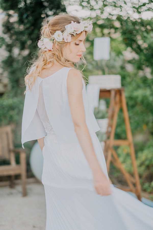 A bridal portrait shot outdoors in a garden setting, showing the bride from behind at a three-quarter angle. She wears a white two-piece bridal outfit with a flowing pleated skirt and a draped cape-style top with flutter sleeves, accented with lace detailing at the waist. Her loose, wavy blonde hair is adorned with a flower crown made of blush pink and ivory blooms. In the soft-focus background, a wooden ladder display holds white framed items, and a wooden chair is visible to the left, suggesting a boho-styled outdoor wedding setup.