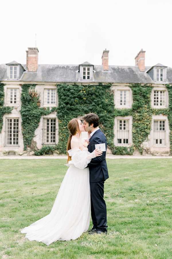 A couple shares a kiss during outdoor wedding portraits on the grounds of a French chateau, with the ivy-covered stone facade of the building prominently visible behind them. The bride wears an off-shoulder white gown with voluminous tulle sleeves and a flowing skirt, while the groom is dressed in a dark navy suit; he holds what appears to be a small card or booklet in one hand. The setting is classic French countryside architecture — a multi-story limestone manor with dormer windows, brick chimneys, and dense ivy coverage across the facade. The shot is a medium full-length portrait with the couple centered in the frame against the expansive chateau backdrop. Potential venue feature image.