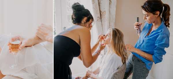 A diptych of two getting-ready images shot indoors in a bright, naturally lit room. The left panel is a close-up detail shot of a bride in a white dress holding a glass of rosé wine. The right panel shows the bride seated and being attended to by two women: one in a black strapless top applying makeup or adjusting her earring, and another in a bright blue button-up shirt holding a makeup brush or tool, likely a hair and makeup artist. The bride is wearing a white robe or wrap top and has long blonde hair. The setting appears to be a preparation room with sheer curtains in the background, and the overall styling is relaxed and candid.