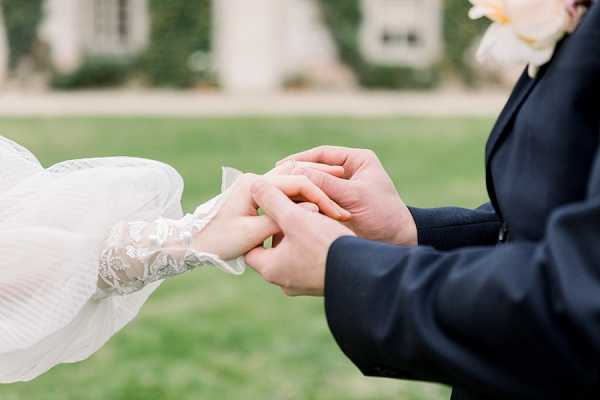 Close-up detail shot of a ring exchange moment during an outdoor ceremony, with the groom in a navy suit placing a ring on the bride's finger. The bride wears a white dress with long lace sleeves featuring delicate floral detailing, and a peach or pale coral flower boutonniere is visible on the groom's lapel. The background is softly blurred, showing a lawn and what appears to be a French chateau or manor house facade with ivy-covered walls. The composition is tightly framed on the couple's hands, keeping the focus entirely on the ring exchange.