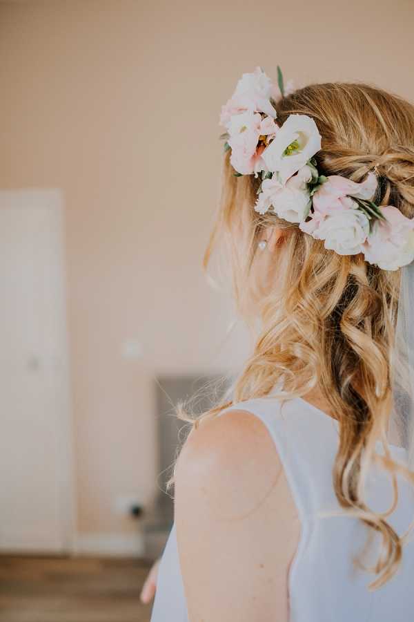 A close-up portrait of a bride taken from behind during the getting-ready phase, shot indoors in a simply furnished room with neutral beige walls. The bride has blonde wavy hair styled in a half-up, half-down arrangement with loose curls, adorned with a floral crown featuring blush pink and ivory lisianthus blooms with small green leaves. She is wearing a white sleeveless dress, visible from the upper back. The composition focuses on the hair and floral accessory detail, with a softly blurred background.
