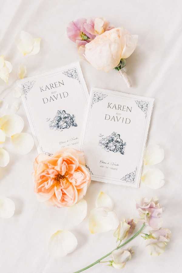 A flat lay detail shot of two matching white wedding ceremony booklets printed with 'Karen and David' in black serif typography, featuring a vintage-style illustrated vignette and ornate corner borders. The booklets are arranged on a white fabric surface surrounded by scattered ivory petals, a peach garden rose, blush sweet peas, a mauve sweet pea stem, and a small boutonniere made of a blush peony bud paired with a pale pink sweet pea. The stationery design follows a classic French-inspired aesthetic with navy ink illustration and references 'Château de Varède' as the venue. The overall color palette is soft blush, peach, and ivory.