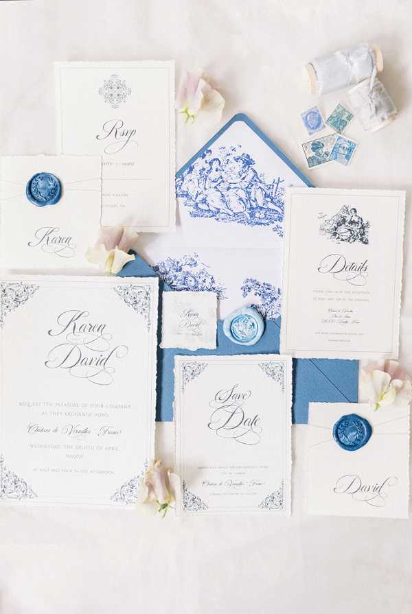 A flat lay of a complete wedding stationery suite for Karen and David, set against a white linen background. The collection includes a main invitation, RSVP card, details card, save the date, envelope liners, and place cards, all featuring deckled (torn) edges and calligraphy script typography in dusty blue and navy ink. The envelope liner is printed with a blue-and-white toile de Jouy pattern depicting pastoral scenes, and the suite is sealed with powder blue wax seals embossed with a crest motif. Scattered around the stationery are loose blush and white sweet pea blooms, vintage blue postage stamps, and a spool of pale blue silk ribbon, completing the French-inspired blue-and-white palette.