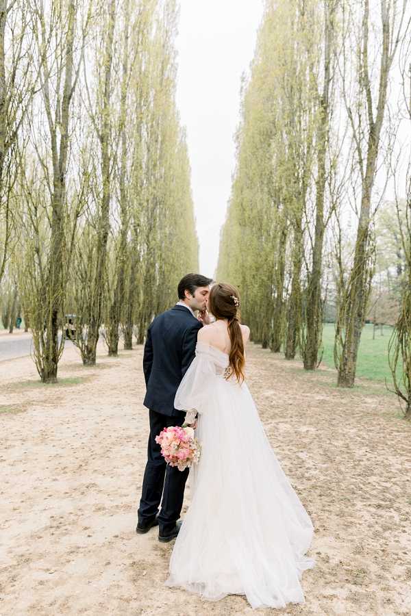A couple portrait taken outdoors along a formal tree-lined sandy avenue of tall poplar trees. The bride and groom are kissing at the center of the frame, positioned mid-distance from the camera in a wide portrait shot. The groom wears a dark navy suit, while the bride wears an off-the-shoulder white ballgown with a full tulle skirt and a long train. Her hair is styled in a loose low updo with a delicate gold hair accessory. She holds a bouquet of pink and coral peonies or garden roses. The composition uses the symmetrical avenue of trees as a natural leading line framing the couple.