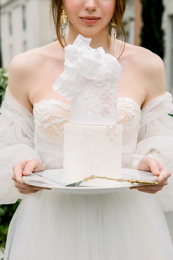A close-up portrait shot of a bride holding a two-tier ivory wedding cake on a white plate, accompanied by a gold-handled cake server and knife. The cake features delicate lace-textured fondant detailing and is topped with a large oversized white sugar or wafer paper flower. The bride is wearing an off-shoulder ivory gown with sheer sleeves and floral lace appliqué detailing, paired with gold drop earrings. The composition is cropped from the chin down, centering the cake as the focal point against a softly blurred outdoor background.