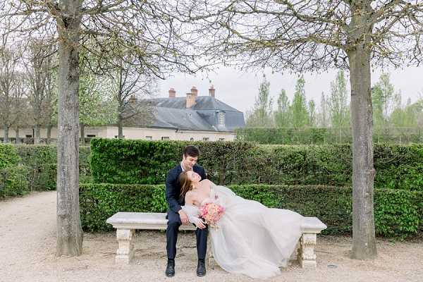A couple portrait taken outdoors in a formal French garden, with the bride and groom seated together on a stone bench between two bare-branched trees. The groom wears a navy suit and the bride wears a full-skirted white gown with a flowing tulle skirt; she holds a bouquet of blush and coral pink flowers. They are leaning toward each other in an intimate pose, framed by a neatly trimmed green hedge behind them, with a classic French chateau building visible in the background. The shot is a wide portrait composition taken in natural, overcast light.