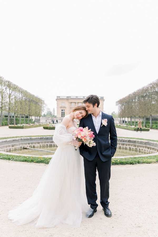 A couple portrait taken outdoors in a formal French garden, with a classical stone pavilion building visible in the background flanked by symmetrically trimmed hedgerows and rows of bare trees. The bride wears an off-the-shoulder white ballgown with voluminous tulle skirt and long sheer puff sleeves, and she leans her head against the groom's shoulder while holding a bouquet of coral, pink, and blush blooms including what appear to be garden roses and sweet peas. The groom wears a dark navy suit with an open-collar white shirt and a small blush boutonniere, with one hand in his pocket. A circular stone fountain basin is visible directly behind the couple. The shot is a full-length portrait with soft, even natural light and a light, airy color palette. Potential venue feature image.