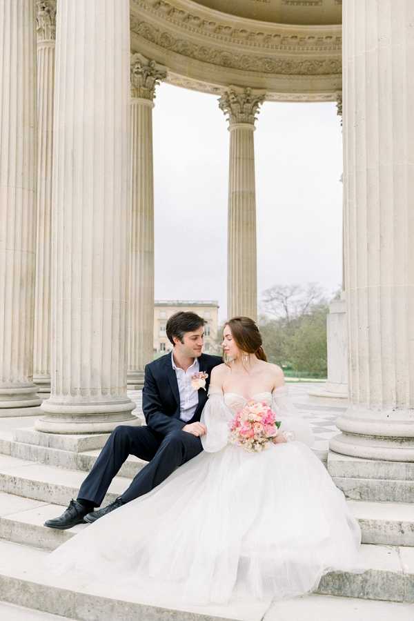 A couple sits together on the stone steps of a neoclassical rotunda or colonnade, surrounded by tall Corinthian columns. The bride wears an off-the-shoulder white ballgown with voluminous tulle skirt and detachable puff sleeves, holding a bouquet of coral, blush pink, and peach blooms including what appear to be peonies and ranunculus. The groom is dressed in a navy suit with a light pink boutonniere, and the two are facing each other in an intimate portrait moment. The shot is a medium-wide portrait with a classic, formal styling aesthetic that complements the ornate architectural setting.