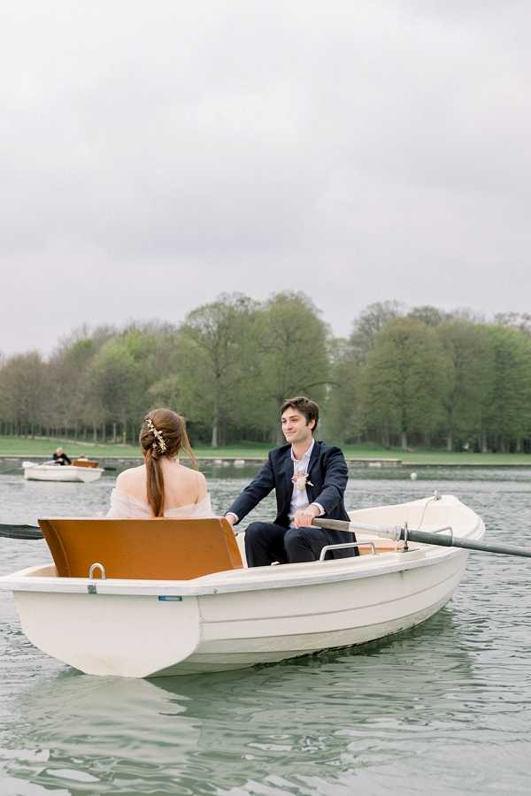 A couple portrait taken on a lake, with the bride and groom seated in a small white and tan wooden rowboat on calm water. The groom, wearing a navy suit, holds the oars and faces the camera with a relaxed expression, while the bride sits with her back to the camera, wearing a sheer long-sleeve dress and an updo hairstyle adorned with a delicate gold floral hair accessory. A second rowboat with two passengers is visible in the background. The setting appears to be a formal park or château grounds, with rows of early-spring trees lining the far bank. The shot is a medium portrait taken from slightly above water level, with an overcast, diffused light.