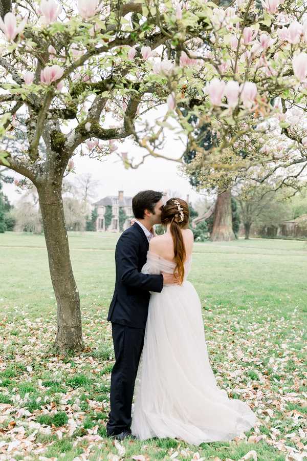 A couple shares a kiss outdoors on the grounds of a French country estate or manor house, visible in the background. The groom wears a dark navy suit, while the bride wears an off-the-shoulder blush-toned tulle ballgown with a flowing train; her auburn hair is styled in a low ponytail adorned with a small floral hair accessory. They are standing beneath a blooming magnolia tree with large pale pink blossoms, and fallen petals are scattered across the lawn around them. The portrait is shot at a medium distance, framing the couple centrally with the magnolia branches arching overhead and the manor's facade softly out of focus in the background.
