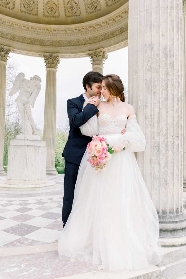 A couple portrait shot outdoors at a neoclassical rotunda or temple structure featuring tall fluted columns, an ornately decorated domed ceiling, a marble checkered floor, and a white marble angel sculpture visible in the background. The bride wears a white strapless tulle ballgown with detachable off-the-shoulder puff sleeves and holds a rounded bouquet of coral and pink peonies with light green foliage. The groom wears a navy suit and leans in to nuzzle the bride's cheek while embracing her from behind. The composition is a medium full-length portrait with a classic, romantic styling aesthetic. Potential venue feature image.