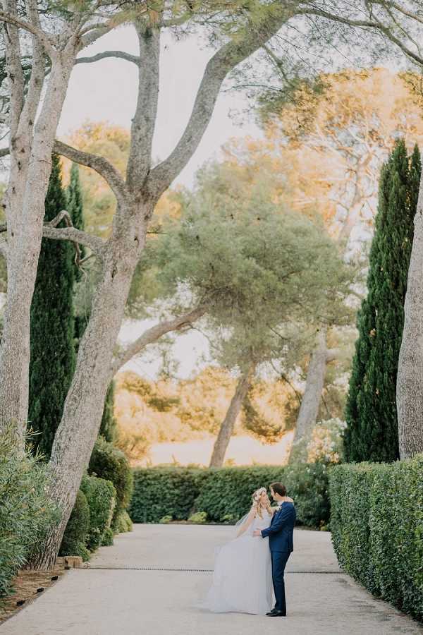 A bride and groom share a close embrace on a formal garden pathway lined with neatly trimmed hedges and tall cypress trees, with large umbrella pines framing the scene. The bride wears a full-length white ball gown with a long veil and a floral crown in her hair, while the groom is dressed in a navy blue suit. The couple is positioned small within the frame, emphasizing the scale and depth of the formal garden alley stretching behind them. This is a wide portrait shot taken outdoors during golden hour light, with a classic French garden setting.