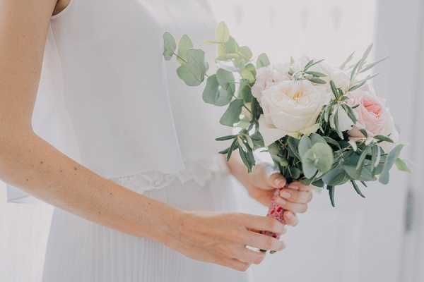A close-up detail shot of a bride holding a small bridal bouquet against her white dress. The bouquet features cream garden roses, blush pink roses, and mixed eucalyptus and olive foliage, with the stems wrapped in pink ribbon. The bride is wearing a white dress with a lace or embroidered cap sleeve detail visible at the shoulder. The overall palette is soft white, blush pink, and sage green.