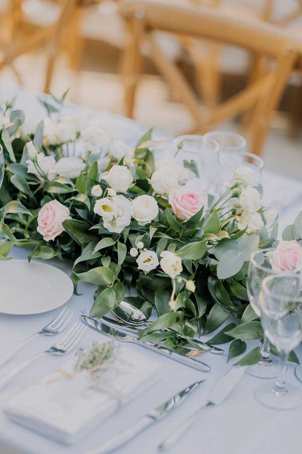 Close-up detail shot of a reception table set with a lush floral runner as the centerpiece, featuring blush pink roses, ivory lisianthus, and abundant green foliage arranged along a white linen tablecloth. Place settings include white plates, silver cutlery, clear glassware, and small folded white place cards tied with a ribbon. Natural crossback wooden chairs are visible in the soft-focus background, suggesting an outdoor or covered terrace setting. The overall decor palette is white, blush pink, and green, consistent with a classic, garden-inspired styling.