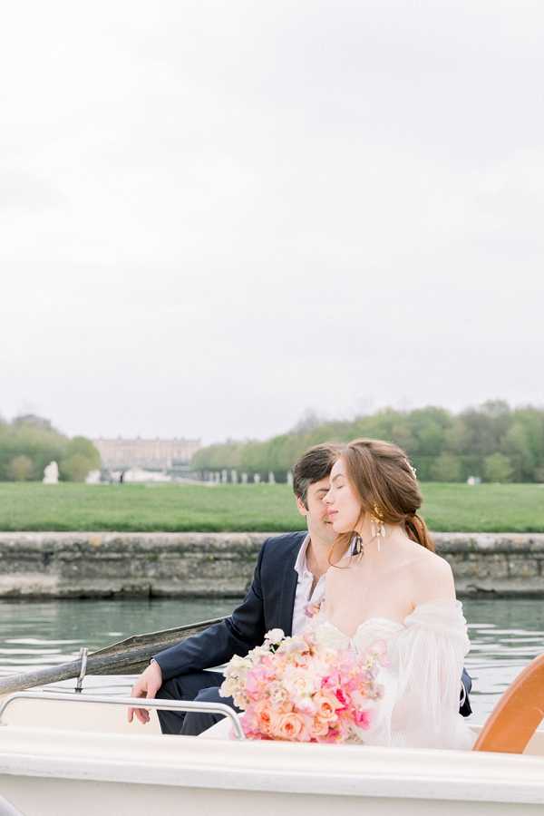 A couple portrait taken on a small boat on a canal or lake, with what appears to be the Palace of Versailles and its formal gardens visible in the background. The bride wears an off-the-shoulder white gown with billowing sleeves and holds a lush bouquet of peach garden roses, blush pink blooms, and bright pink flowers. The groom is dressed in a navy suit with an open-collar shirt, and the two lean into each other with eyes closed in a tender moment. The composition is a mid-range portrait shot with soft, diffused natural light giving the image a pale, airy tone.