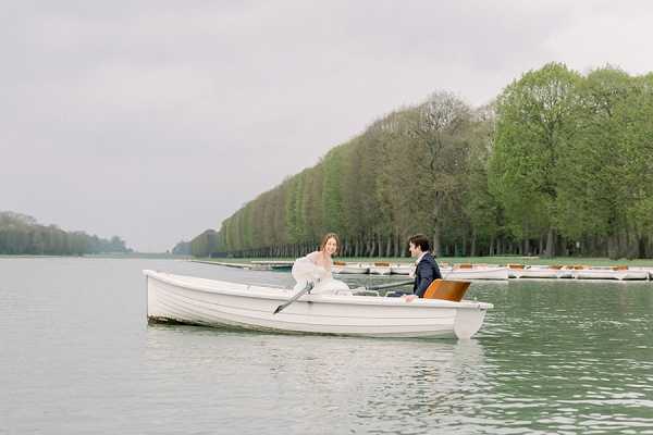 A bride and groom sit facing each other in a small white rowboat on a calm lake, appearing to share a conversation. The bride wears a white gown and the groom is dressed in a dark navy suit. Several additional white and wood-trimmed boats are docked along the shore in the background, flanked by a formal row of neatly trimmed trees typical of French formal garden design. The image is a wide shot with soft, overcast light giving the scene a muted, cool palette. The setting appears to be the Grand Canal at the Palace of Versailles.