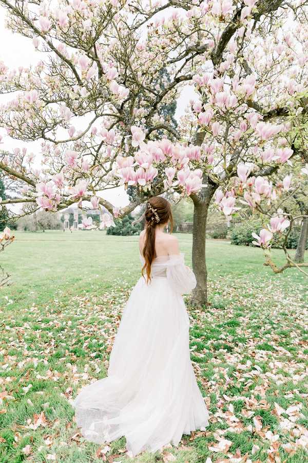 A bridal portrait taken outdoors in the grounds of what appears to be a French country estate or chateau, visible in the soft background. The bride stands with her back to the camera beneath a large flowering magnolia tree covered in pink blooms, with fallen petals scattered across the grass below. She wears an off-the-shoulder white tulle ball gown with a flowing train, and her auburn hair is styled in a half-up ponytail adorned with a delicate floral or crystal hair accessory. The composition is a full-length portrait with a romantic, airy aesthetic using soft natural light and a blush and white color palette.