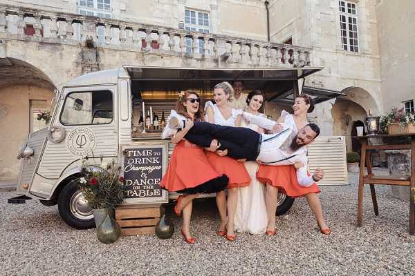 A playful group shot during what appears to be a cocktail hour or reception, set in the gravel courtyard of a French chateau with arched stone architecture visible in the background. The focal point is a vintage cream-colored Citroën HY van converted into a champagne bar, displaying a chalkboard sign reading 'Time to Drink Champagne & Dance on the Table.' The bride wears a white gown and is being carried horizontally by three bridesmaids and the groom, all of whom are laughing; the bridesmaids wear matching coral-orange full skirts with white blouses and coral heels, while the groom is dressed in a navy suit. A wooden crate and vase with wildflowers are positioned beside the van, and a lantern with florals is visible to the right. Medium-wide group shot with a candid, high-energy composition.