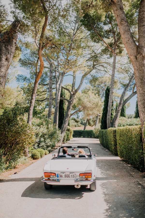 A couple — appearing to be a bride with light curly hair and a groom — ride in a vintage white convertible car (French license plate) along a tree-lined driveway, shot from behind in a wide portrait composition. The setting is an outdoor estate or domaine, with a formal clipped hedge on one side and tall pine trees framing the drive. The styling is classic and relaxed, with the open-top white classic car as a deliberate transport choice. The image is taken from ground level behind the vehicle, capturing the couple from the back as they drive away.