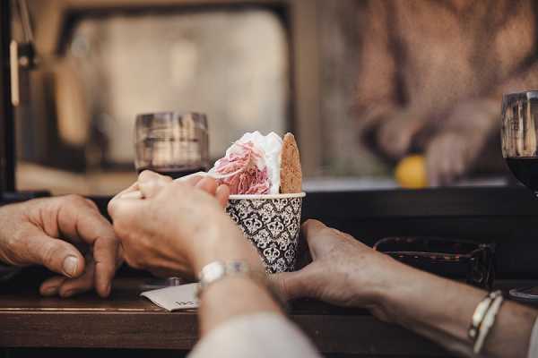Close-up detail shot of a guest receiving an ice cream cone at what appears to be a wedding cocktail hour or reception. The cone is wrapped in a black and white damask-patterned paper sleeve and topped with a scoop of pink ice cream. Two sets of hands are visible — one handing over the cone, one receiving it — with a silver bracelet visible on one wrist. In the background, a glass of red wine and a second dark glass are softly out of focus, suggesting a food and drink station or mobile dessert cart. The styling is relaxed and festive, consistent with a casual outdoor catering setup.