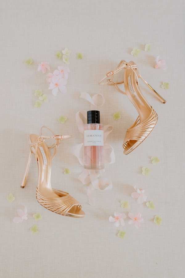 A flat lay detail shot of bridal accessories arranged on a neutral linen-textured cream background, scattered with small pink and pale green florettes and white petals. The central items are a pair of rose gold strappy stiletto heels with multi-strap toe detailing and ankle buckle closures, flanking a bottle of Christian Dior 'Dioramour' perfume with a blush pink liquid and black cap. The overall color palette is soft rose gold, blush pink, and cream, consistent with a classic or modern romantic bridal styling theme.