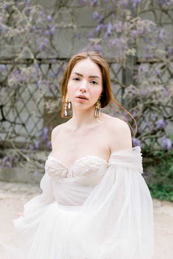 A close-up portrait of a bride standing outdoors against a wall covered in blooming wisteria with soft purple clusters. She wears an off-the-shoulder ivory wedding gown with a lace-embroidered sweetheart bodice and flowing, billowing tulle sleeves and skirt. Her accessories include statement gold chandelier earrings with pearl or crystal drops. Her auburn hair is loosely styled and slightly windswept. The composition is a medium portrait shot with the bride as the sole subject, styled in a romantic, soft aesthetic with a light, airy color palette of ivory and muted lavender.