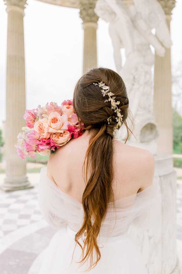A close-up portrait of a bride shot from behind, showcasing her hair and bouquet against a classical architectural backdrop featuring white marble columns and a large sculpted statue. The bride wears an off-the-shoulder white gown and her brunette hair is styled in a loose low ponytail with a braided section, adorned with a gold leaf-and-branch hair comb. She holds a bouquet of hot pink sweet peas, peach garden roses, and blush blooms resting over her shoulder. The setting appears to be an outdoor classical temple or colonnade with a black-and-white checkered marble floor, consistent with French formal garden architecture.