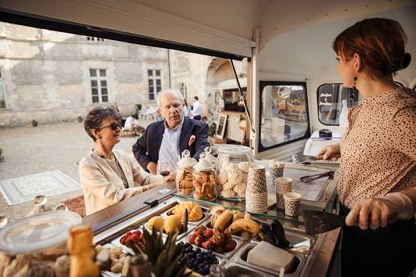 A cocktail hour or reception scene photographed from inside a vintage-style food or dessert cart, showing two wedding guests — a woman in a beige blazer and a man in a navy suit with a pocket square holding a glass of red wine — being served by a staff member in a white polka-dot blouse. The cart's counter displays fresh fruit including strawberries, blueberries, and bananas, along with glass jars filled with biscuits and meringues, and stacked paper cups. In the background, a classic French chateau courtyard is visible with additional guests mingling at tables. The composition is a medium shot taken from the vendor's perspective inside the cart, giving an immersive point-of-view framing. Potential venue feature image.
