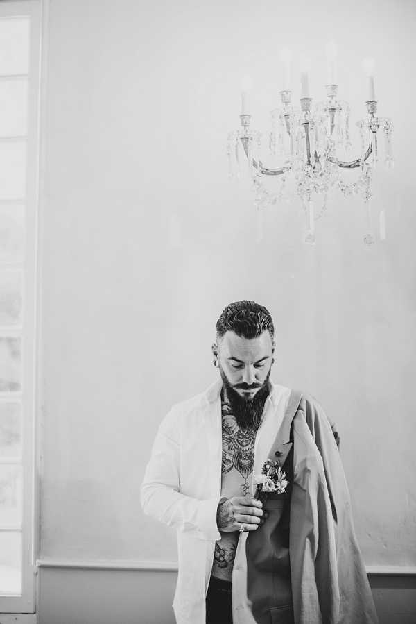 A black-and-white getting-ready portrait of the groom indoors, standing against a light-toned wall beneath a crystal candelabra chandelier. The groom has a full beard, slicked-back hair, large ear gauges, and extensive chest and arm tattoos visible through his open white dress shirt; he holds his suit jacket over one arm and looks down at a boutonniere he is pinning or adjusting. The image is a mid-length portrait with soft, high-key tones and gentle contrast, giving the room a bright, airy feel.