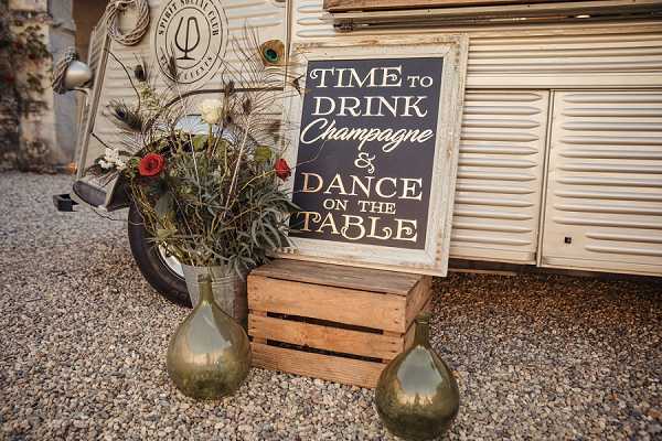 A detail shot of a rustic wedding reception vignette set on a gravel surface in front of a vintage cream-colored caravan or food truck. A chalkboard-style sign with a distressed white frame reads 'Time to Drink Champagne & Dance on the Table' in gold and white lettering, propped against a weathered wooden crate. Two brass or antique gold teardrop-shaped vases flank the crate, and a third larger brass vase holds a wild, loosely arranged bouquet of red flowers, dried grasses, eucalyptus, and feathery foliage. The overall styling is bohemian-rustic with a warm metallic and natural palette.