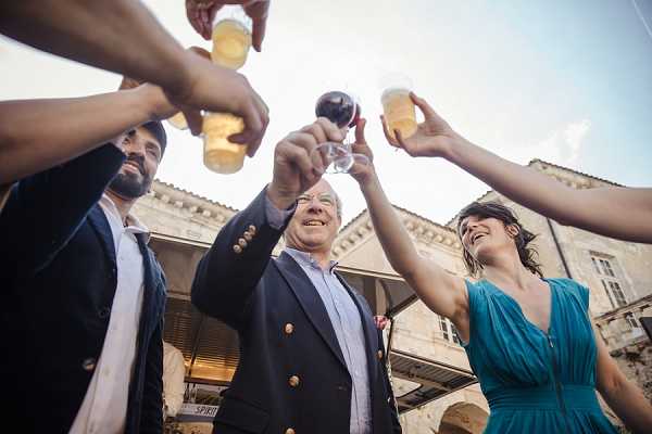 A group of wedding guests raises glasses in a toast during what appears to be a cocktail hour or reception, photographed from a low angle looking upward. At least four people are visible, including a man in a navy blazer with gold buttons at center smiling broadly, a man in a dark suit to his left, and a woman in a teal draped dress to his right. Drinks include what appear to be glasses of white wine, beer bottles, and a glass of red wine. The setting is an outdoor courtyard of a historic stone building with arched architectural details visible in the background. The low-angle composition emphasizes the raised glasses converging toward the center of the frame.