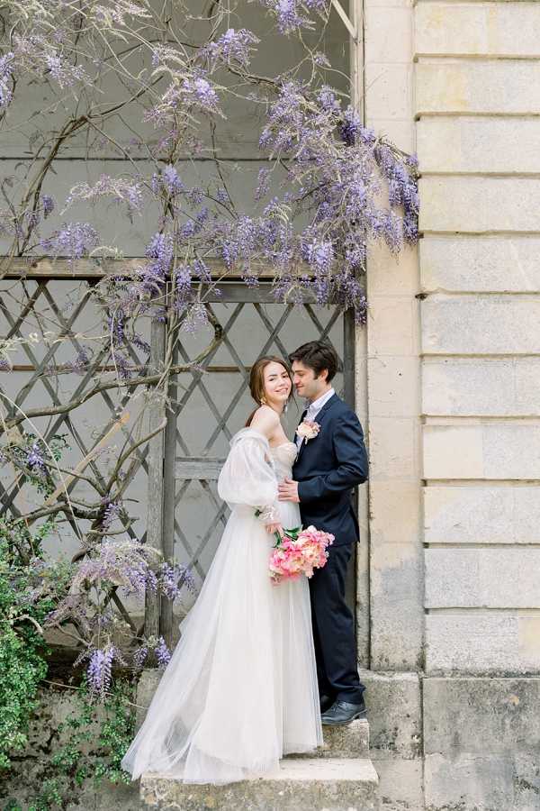A couple portrait taken outdoors against the stone facade of a French chateau or historic building, with blooming wisteria cascading overhead and a latticed wooden door in the background. The bride wears an off-the-shoulder white tulle ballgown with voluminous sheer puff sleeves and holds a bouquet of coral and pink peonies; the groom wears a navy suit with a blush boutonniere. The two are close together, the groom nuzzling the bride's cheek as she smiles with her eyes closed. The image is a medium full-length portrait with soft, even natural light.