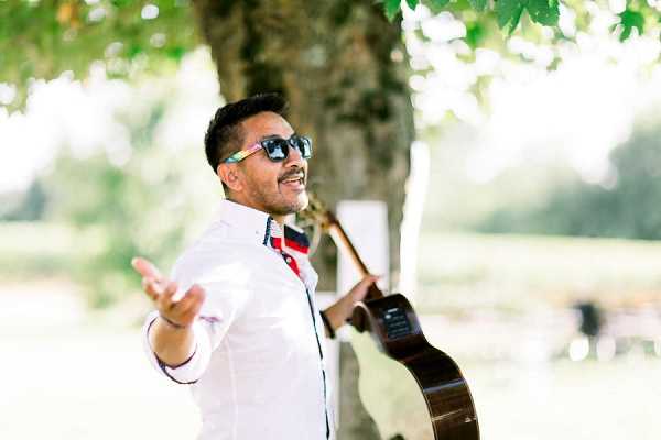 A male musician performs outdoors at what appears to be a wedding cocktail hour or reception, holding an acoustic guitar in one hand while extending his other hand outward in an expressive gesture. He is wearing a white button-down shirt with a red and navy collar detail and dark sunglasses, and appears to be singing or engaging with guests. The setting is an outdoor garden or grounds with a large tree in the background. The shot is a mid-length portrait with a shallow depth of field, keeping the performer sharp against a softly blurred green outdoor background.