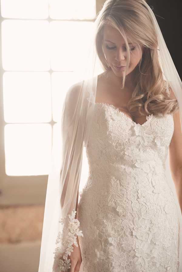A bridal portrait taken indoors, with the bride positioned in front of a large window that creates strong backlighting and a warm, overexposed glow. The bride wears a fitted ivory strapless lace gown with an allover floral lace pattern and a sweetheart neckline, paired with a long cathedral-length veil edged with appliqué lace detailing. Her blonde hair is styled in loose curls falling over one shoulder. She is looking downward, and the composition is a close-up portrait cropped at the waist, emphasizing the texture of the lace dress and veil.