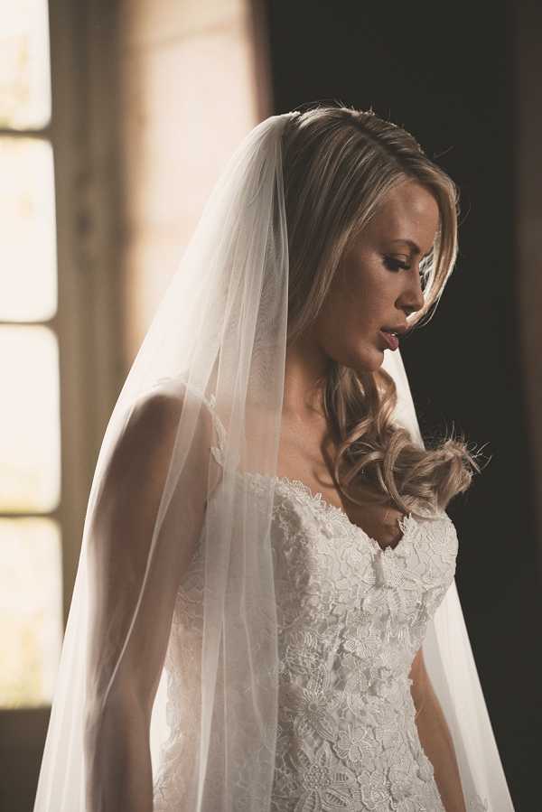 A close-up bridal portrait of a single bride looking downward, shot indoors with warm, directional natural light coming from a window behind her. She wears an ivory strapless lace wedding gown with a detailed floral lace bodice and a long, plain-edged cathedral or elbow-length ivory veil draped over her shoulders. Her blonde hair is styled in soft, loose curls worn to one side. The portrait is tightly framed from approximately the waist up, with a dark background contrasting against the warm window light to create strong shadow and highlight contrast on her face and dress.