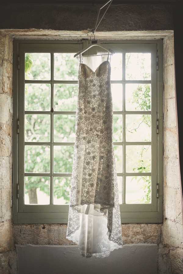 A getting-ready detail shot of a wedding dress hanging on a hanger in front of a multi-pane window with aged sage-green painted frames set into a rustic stone wall. The gown is a strapless, sweetheart-neckline silhouette with an overlay of ivory and taupe floral lace over what appears to be a champagne-toned underlining, giving it a muted, antique appearance. The lace hem has a slight train that pools at the base of the window sill, where a pair of white platform bridal shoes is also visible. Natural backlight from the window illuminates the lace pattern, and the overall image has a soft, slightly warm, faded tone consistent with a film-inspired edit.
