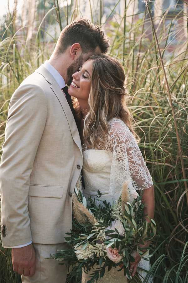 A couple portrait shot outdoors, with the groom nuzzling into the bride's temple as she smiles with her eyes closed. The groom wears a light sand-beige linen suit with a white shirt and dark tie, while the bride wears a white lace wedding dress featuring sheer three-quarter-length sleeves with floral lace detailing. The bride holds a large, loosely arranged bouquet incorporating greenery and olive branches, blush pink roses, pampas grass, and dried neutral-toned florals, consistent with a boho styling aesthetic. The portrait is a medium close-up framed against tall ornamental grasses that fill the background, with warm golden-hour light filtering through.