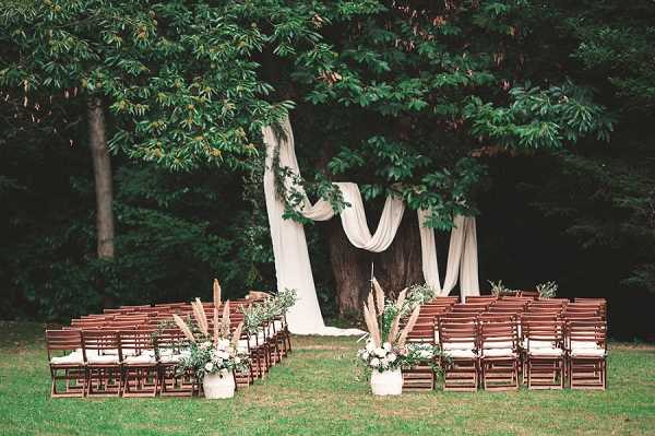 An outdoor wedding ceremony setup in a garden or wooded area, with no guests or couple present. The altar is formed by a large tree trunk draped with flowing ivory fabric panels and greenery vines, creating a natural boho-style backdrop. Rows of dark wood folding chairs with white seat cushions are arranged in two sections facing the altar, accommodating approximately 60–80 guests. At the base of the aisle and flanking the altar, white ceramic pots hold arrangements of ivory and white florals, dried pampas grass, and greenery, consistent with a natural, earthy boho styling palette. Wide shot composition capturing the full ceremony setup.