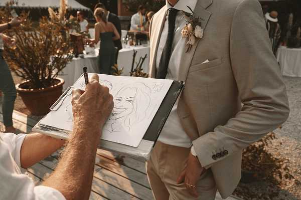 A caricature artist is drawing a portrait sketch on a clipboard during what appears to be an outdoor cocktail hour, with the groom standing nearby as the likely subject. The groom wears a light beige linen suit with a dark tie and a white and cream floral boutonniere. The sketch on the clipboard shows a smiling female face in a caricature style. In the background, guests mingle near white-clothed tables in an outdoor courtyard or terrace setting with terracotta planters visible. The shot is a close-up detail capturing the artist's hand and clipboard in the foreground, with the groom partially visible at center frame.